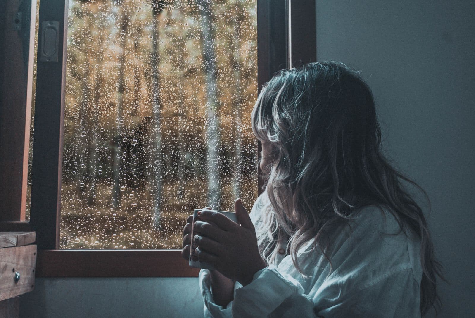 A person looking out a rain-covered window, holding a mug